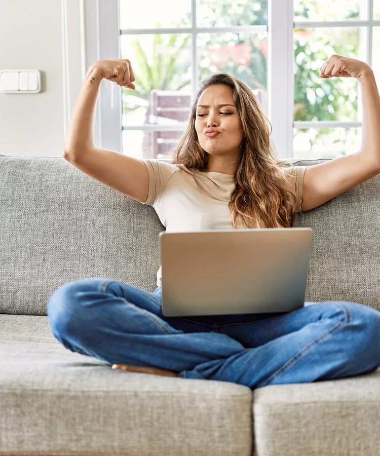 Confident woman celebrating success while working on laptop from home couch flexing arms