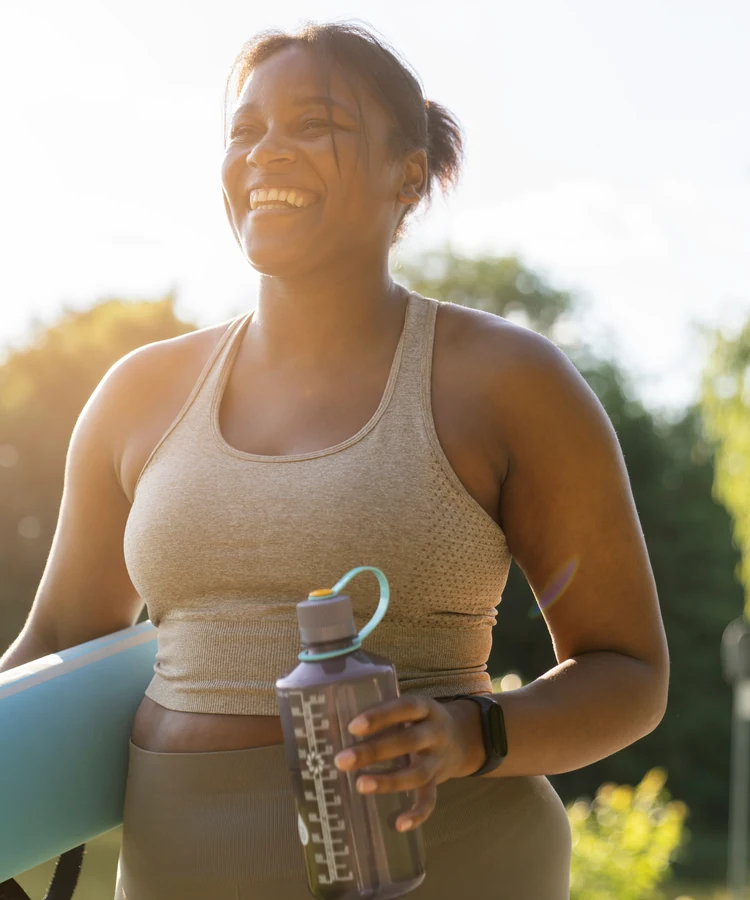 Smiling woman in athletic wear holding yoga mat and water bottle outdoors in natural sunlight for fitness wellness
